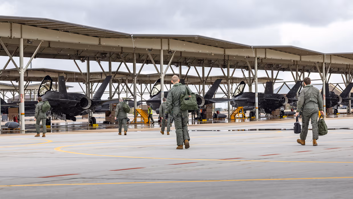 Vliegers lopen met tassen richting F-35’s die onder een overkapping op een platform staan.