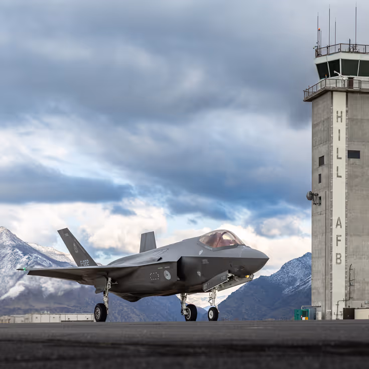 Een F-35 links van de luchtverkeerstoren waarop ‘Hill AFB’ staat. Op de achtergrond besneeuwde bergen.