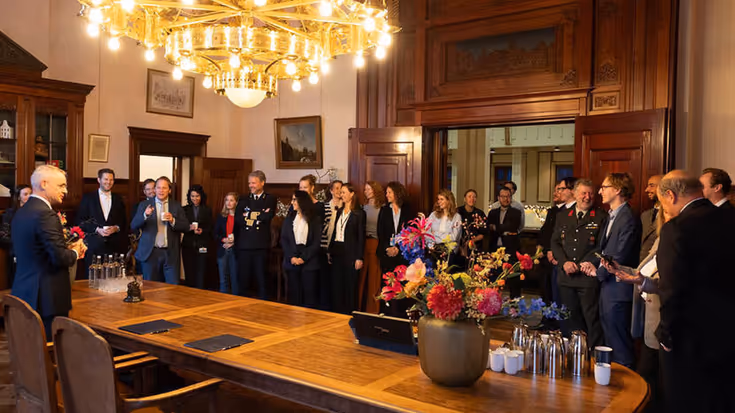Een ovale houten tafel met staande mensen erachter. Boven de tafel een goudkleurige kroonluchter.