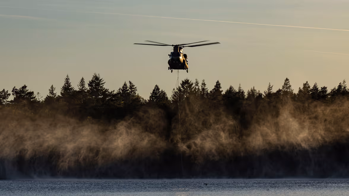 Een Chinook-transporthelikopter hangt laag boven een meer om water te laden in een speciale waterzak.