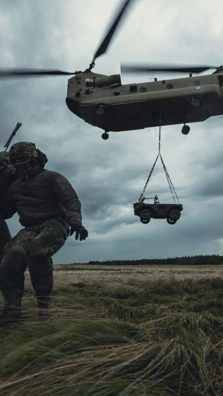 Een Chinook-transporthelikopter stijgt op met een voertuig die eronder hangt. Drie militairen staan op de heide en houden elkaar vast om te blijven staan in het vlieggeweld.