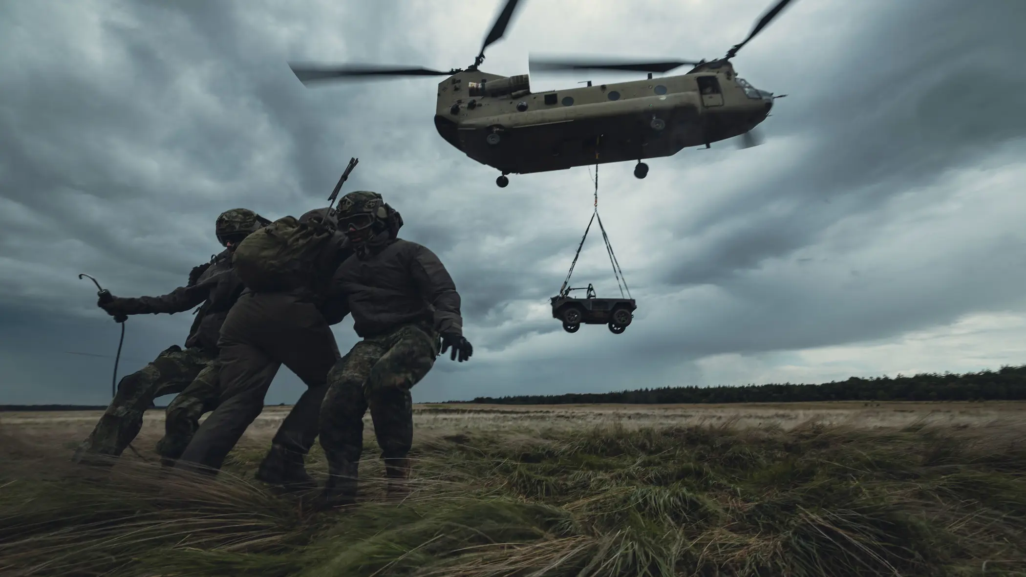 Een Chinook-transporthelikopter stijgt op met een voertuig die eronder hangt. Drie militairen staan op de heide en houden elkaar vast om te blijven staan in het vlieggeweld.