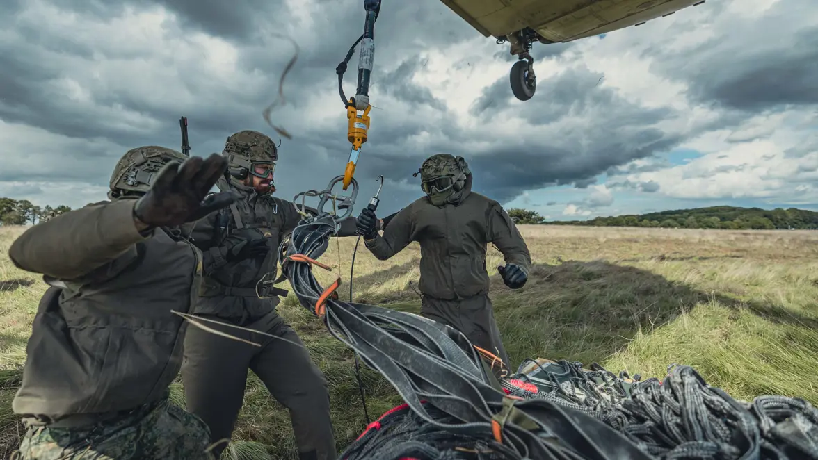 3 militairen staan naast een houten pakket, met daaromheen een net en erboven een haak, die ze vastmaken aan de Chinook die boven hen hangt.