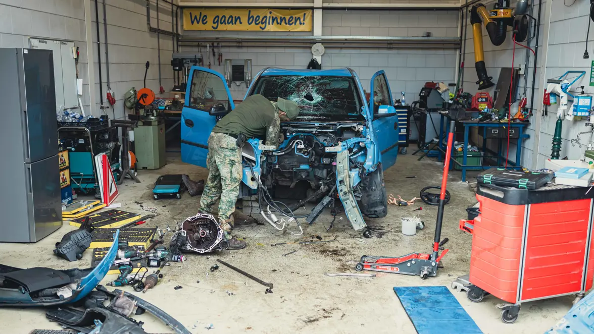 Een militair sleutelt aan een blauw voertuig. Er zit een gat in de voorruit en de auto is volledig beschadigd.