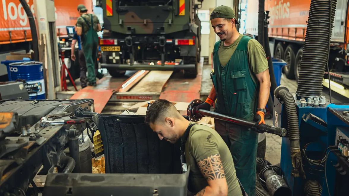 Een militair in een groene overall houdt een buis vast en glimlacht naar een collega die op zijn knieen op de werkplaats zit te sleutelen. 