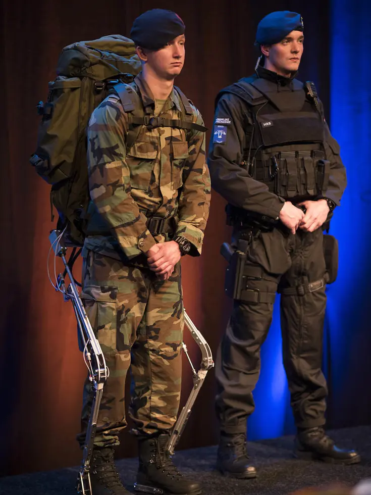 Links: Twee mannen in uniform: één met camouflage, rugzak en exoskelet, de andere in zwart met kogelwerend vest.    Rechts: Een man in grijs jack praat met een soldaat in camouflage en rugzak in een groen bos. 