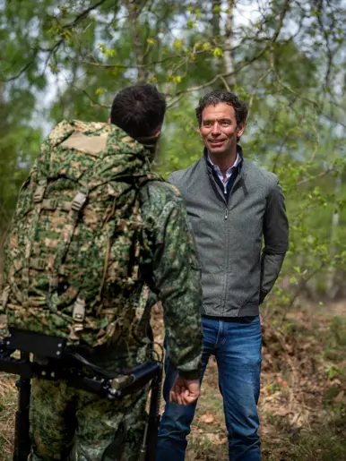 Links: Twee mannen in uniform: één met camouflage, rugzak en exoskelet, de andere in zwart met kogelwerend vest. Rechts: Een man in grijs jack praat met een soldaat in camouflage en rugzak in een groen bos. 