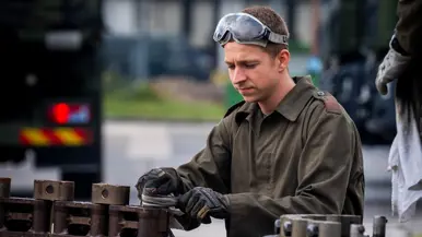 Een militair in overall en met een veiligheidsbril op sleutelt aan een metalen constructie.