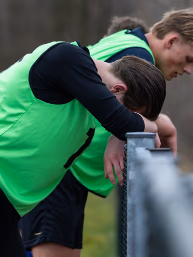 Een groep hardlopers in gele hesjes met nummers rent op een atletiekbaan. De voorste loper lijkt gefocust, met bomen en een hek op de achtergrond.