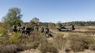 Een groep soldaten in camouflage loopt door een heideveld met lage struiken, een helikopter staat op de achtergrond. 