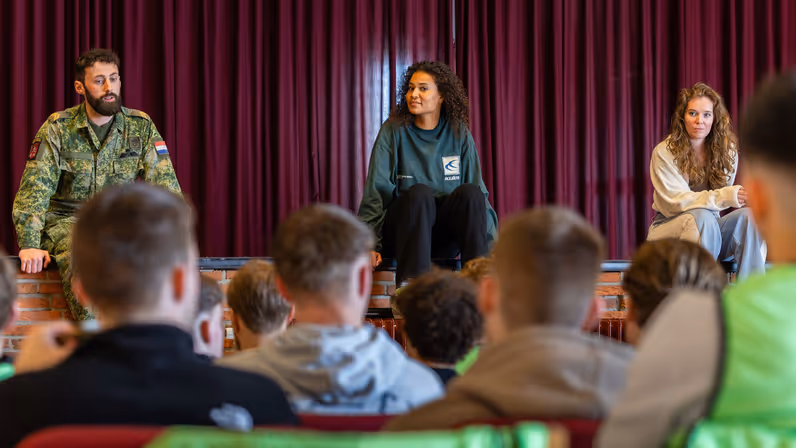 Drie personen zitten op een podium: een militair links, een vrouw in een donkere trui midden, en een vrouw rechts. Een groep jongeren luistert aandachtig in een zaal.