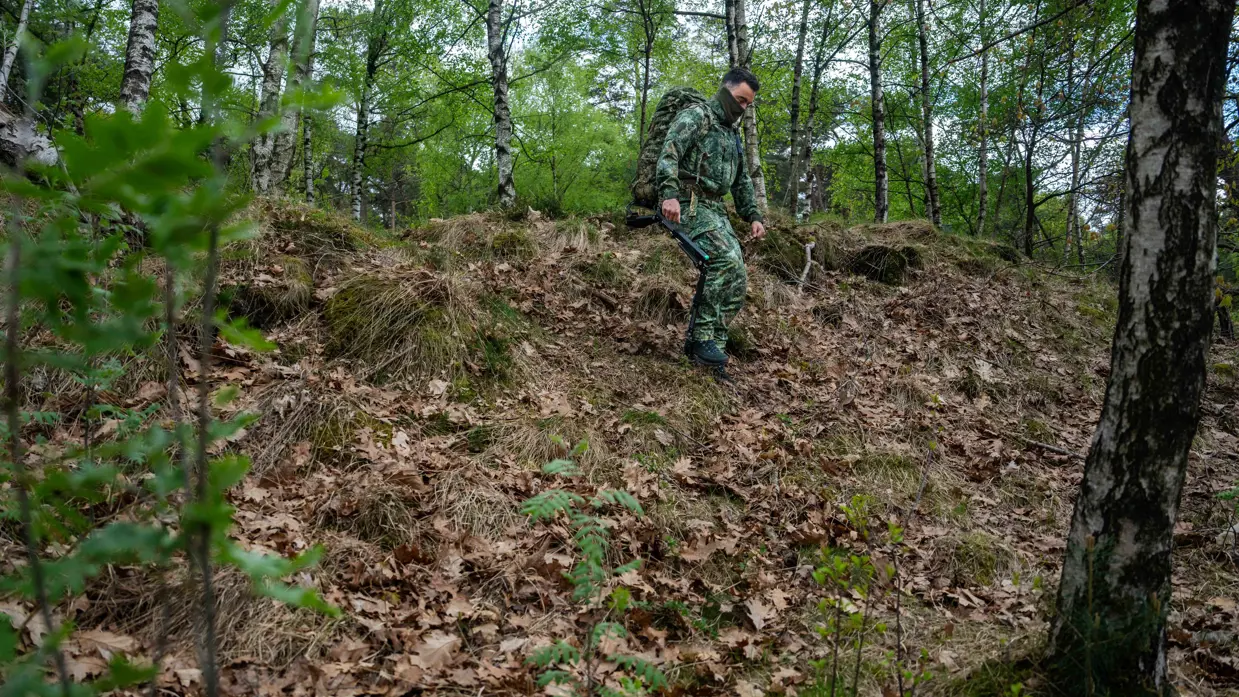 Links: Een soldaat in camouflage met rugzak en exoskelet loopt door een bos met bomen en struiken op een heuvel.    Rechts: een soldaat in camouflage met rugzak en stokken klimt een met bladeren bedekte heuvel op in een groen bos. 