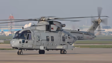  Een grote, grijze militaire helikopter met draaiende rotorbladen staat op een startbaan, met "Amsterdam" en taxiborden op de achtergrond.