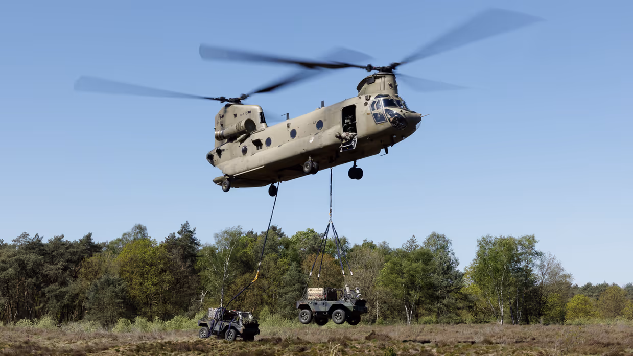 Een militaire helikopter tilt een jeep op boven een bosrijk heideveld, met een andere jeep op de grond. 