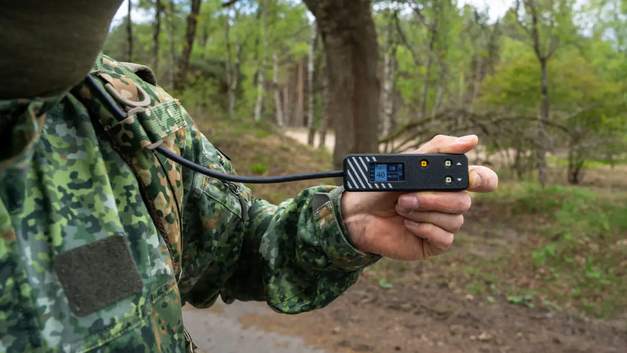 Links: iemand trekt een groene bergschoen aan met een zwarte beugel, buiten op een bosbodem met bladeren.    Midden: Iemand in camouflagebroek zit op een auto met bruine sokken en houdt een groene bergschoen vast. Rechts: Een man in uniform houdt een metertje vast. 