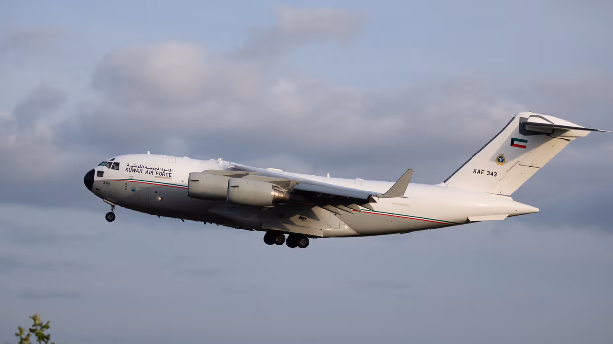Een wit vliegtuig met de tekst "KUWAIT AIR FORCE" en nummer "KAF 343" vliegt door een bewolkte lucht. 