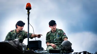 Twee huzaren in camouflage-uniform zitten op een pantservoertuig, de een wijst, de ander luistert onder een oranje lamp.