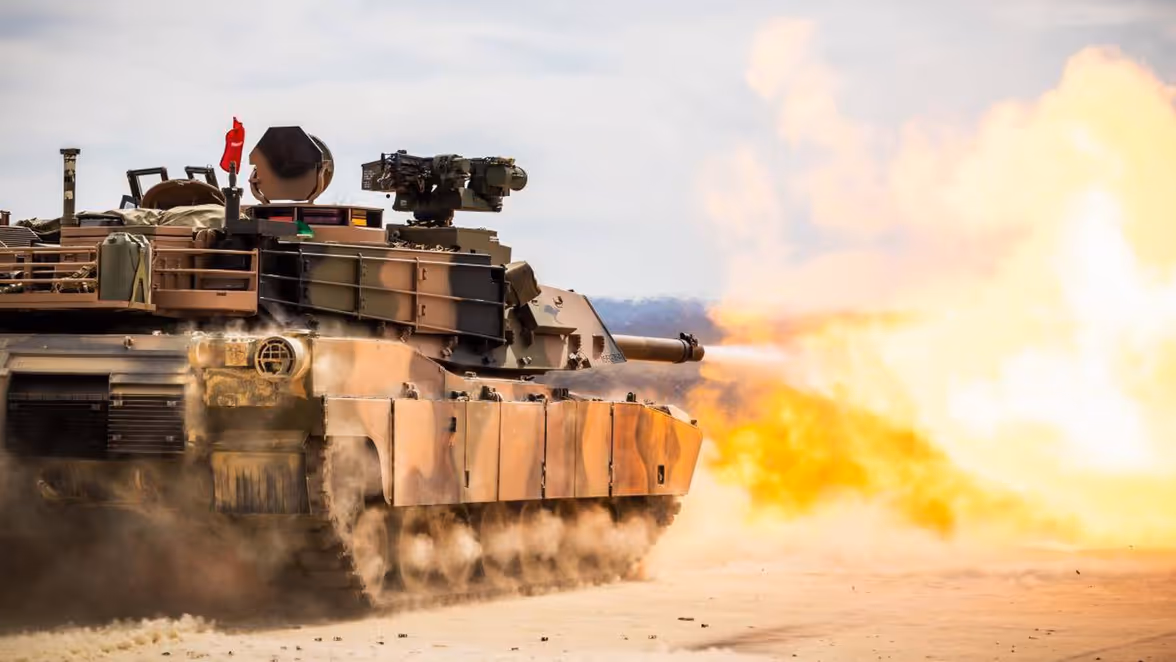 Een tank vuurt zijn kanon af in een woestijn, met zand en vuur dat uit de loop spat, terwijl de tank in beweging is.