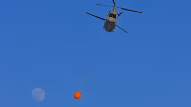 Een helikopter hangt een grote oranje waterzak onder zich, met de volle maan op een diepblauwe hemel. 
