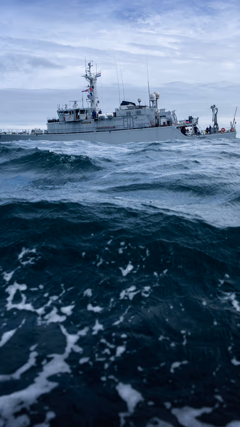 Een groot, grijs schip vaart door woeste, donkerblauwe golven onder een bewolkte, grijze hemel, met witte schuimkoppen op het water.