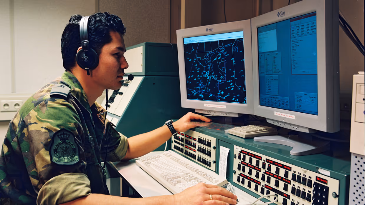 Een militair in camouflagepak achter twee computerschermen. 