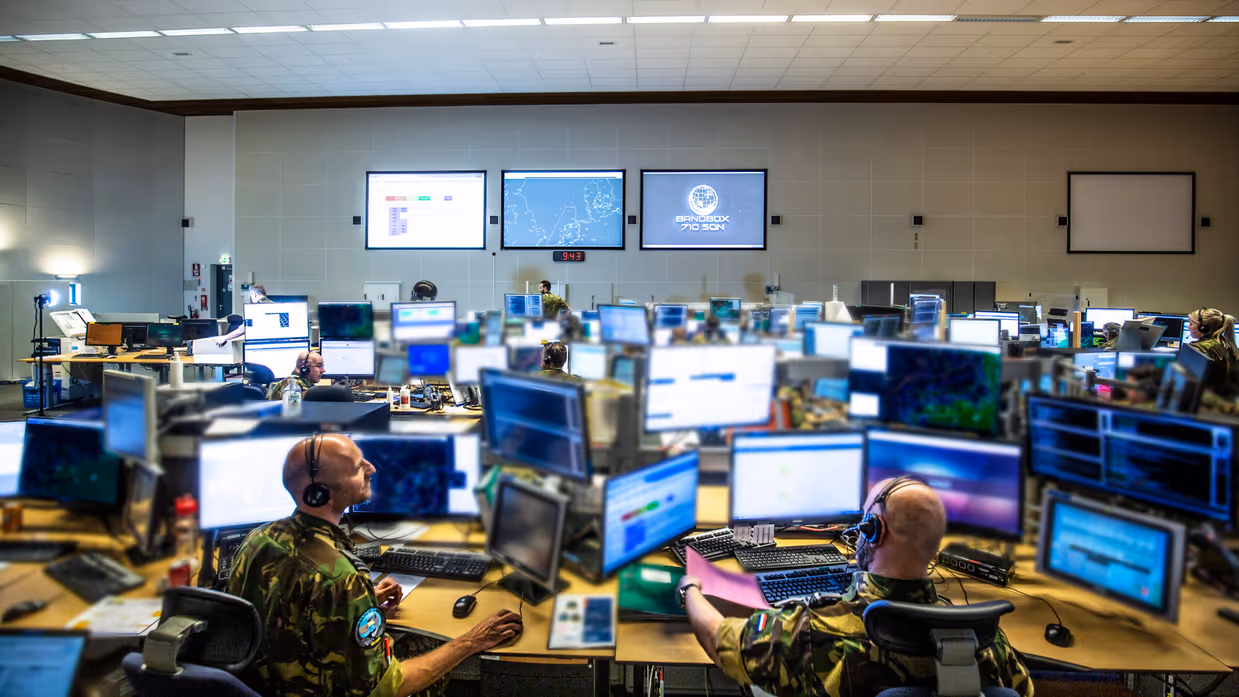 Overzichtsfoto van een ruimte met veel computerschermen. In de achtergrond op de muur hangen drie grote projectieschermen. Foto: 