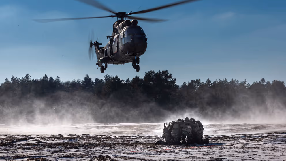 Legerhelikopter hangt net boven de grond terwijl groepje militairen ineengedoken bij elkaar zit om zich te beschermen tegen wind en stof.
