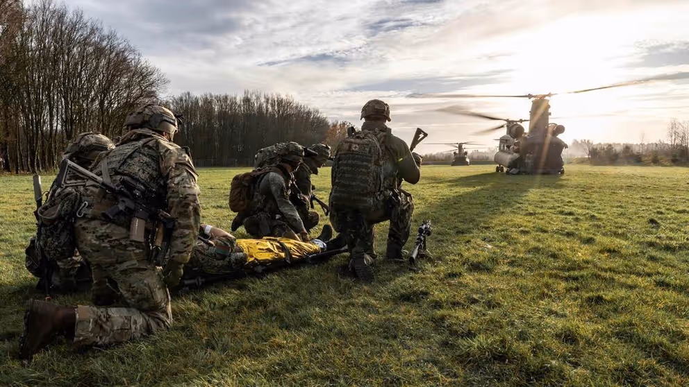 5 militairen zitten op hun knieën in het gras bij een gewonde op een brancard. 2 Chinook-transporthelikopters zijn zichtbaar op de achtergrond.