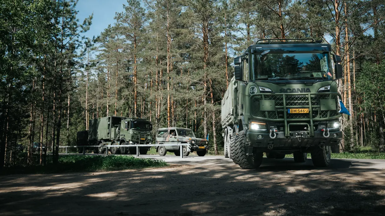 Militaire voertuigen in het bos waarbij een vrachtwagen voorop afslaat richting de camera