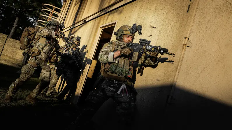 Een booskijkende militair gaat voorwaarts met een lang wapen langs zijn lichaam. Hij en de collega’s achter hem staan op het punt het gebouw rechts van hen binnen te vallen. 