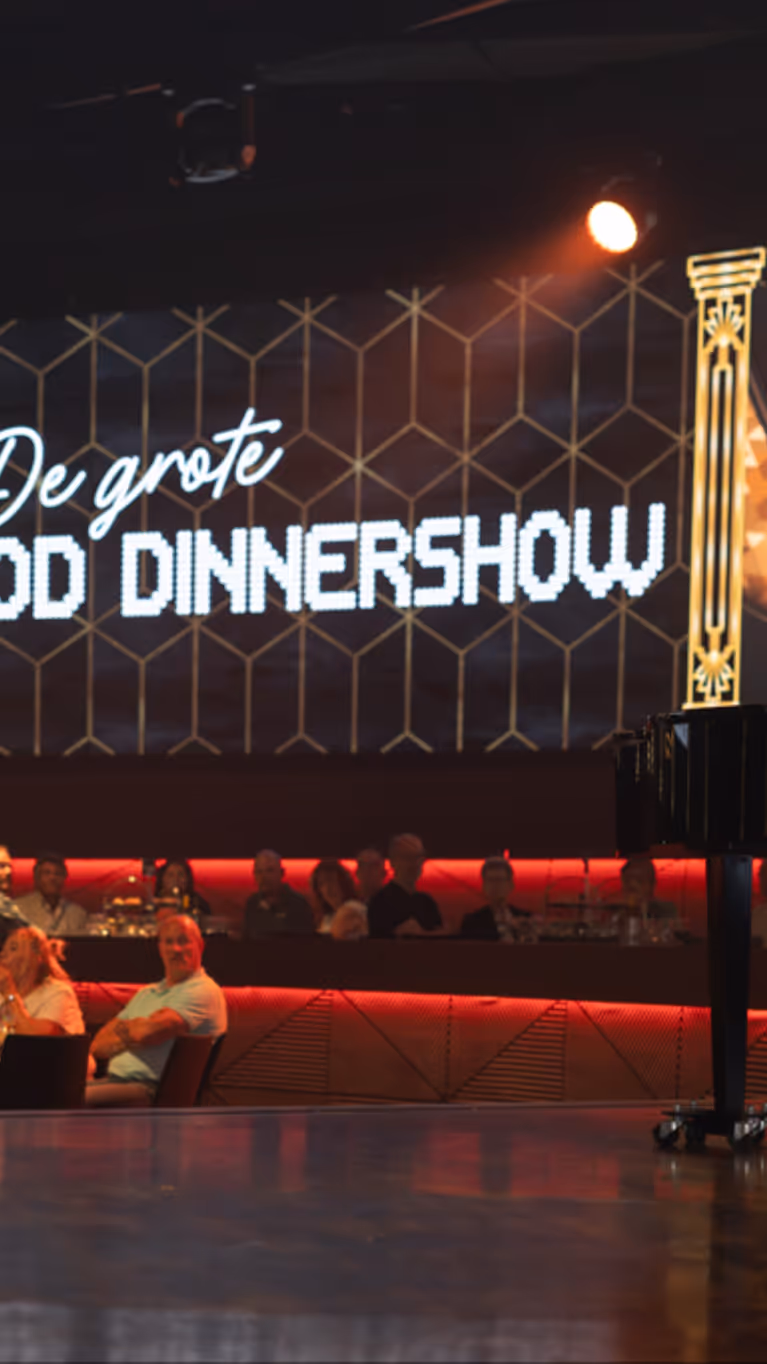 Een zaal vol mensen kijkt naar een man op een podium. Op de achtergrond is een billboard te zien met de tekst ‘De Grote MOD-dinnershow’.