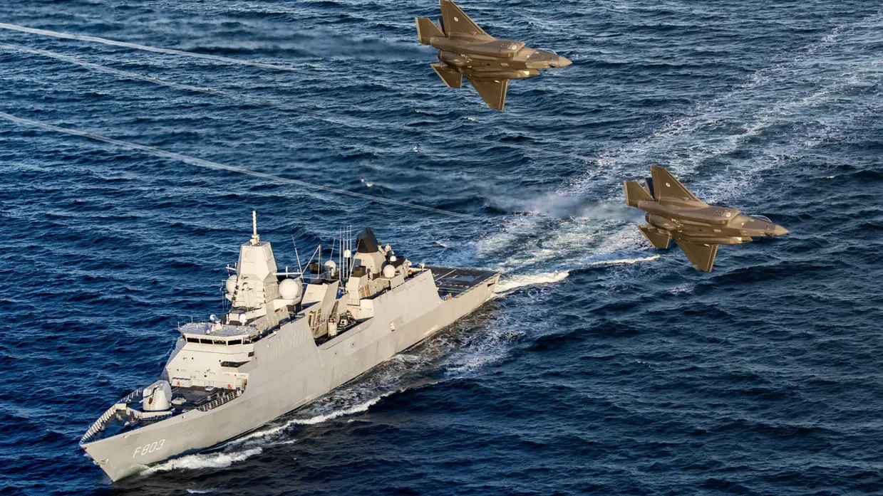 Een marineschip op zee met daarboven 2 gevechtsvliegtuigen.