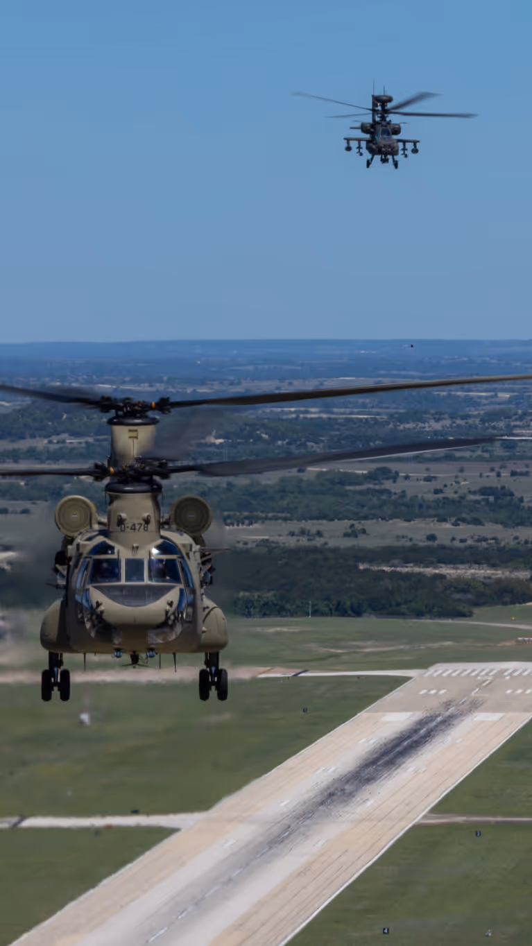 Een Chinook en twee Apaches vliegend boven een startbaan met op de achtergrond een blauwe lucht en weids uitzicht over vlak platteland.