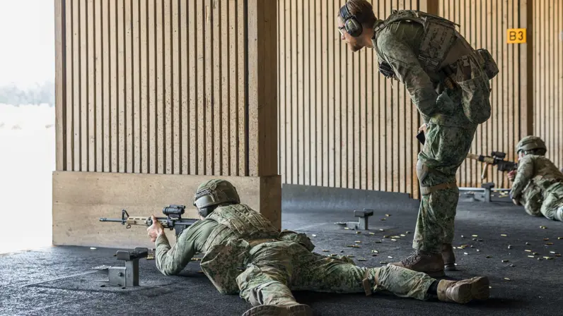 Een militair ligt op zijn buik met een wapen voor zijn gezicht op de schietbaan. Een andere militair staat achter hem en geeft hem instructies.&nbsp;