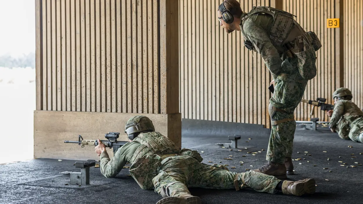 Een militair ligt op zijn buik met een wapen voor zijn gezicht op de schietbaan. Een andere militair staat achter hem en geeft hem instructies.&nbsp;