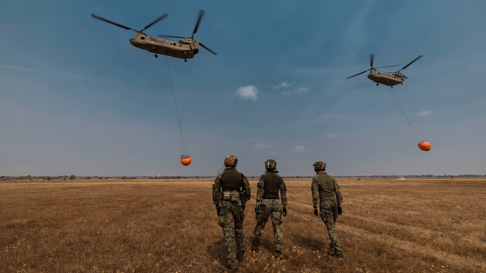 Militairen op de rug gezien. In de lucht 2 helikopters met waterzakken.