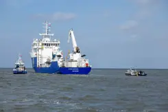 Het schip de NS Geosea, van de onderkant blauw en de bovenkant wit, vaart op zee. Links en rechts een klein bootje.