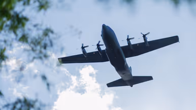 Een C-130 vliegt laag over. Links staan bomen. 