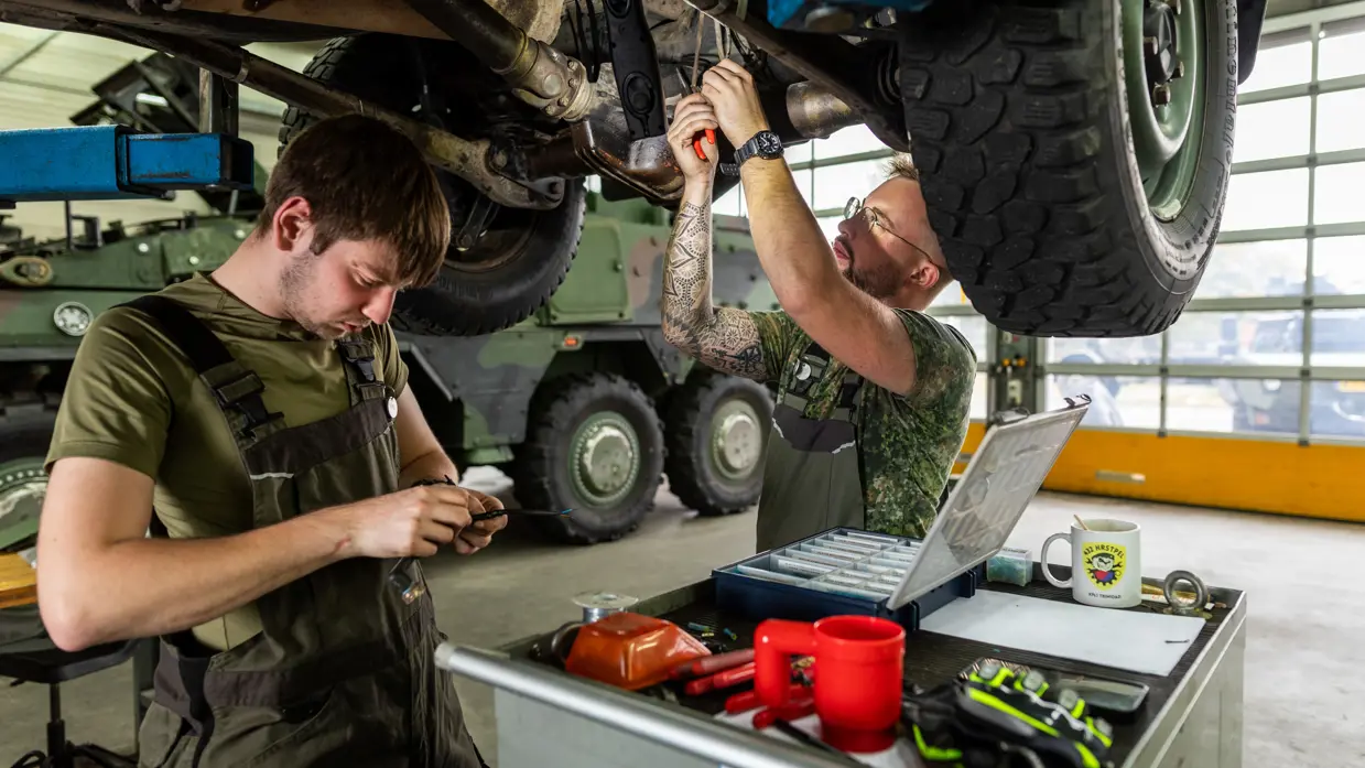 Een man in een groene overall sleutels aan een klein onderdeel in zijn handen. Een ander sleutels boven zijn hoofd aan de onderkant van een voertuig.&nbsp;