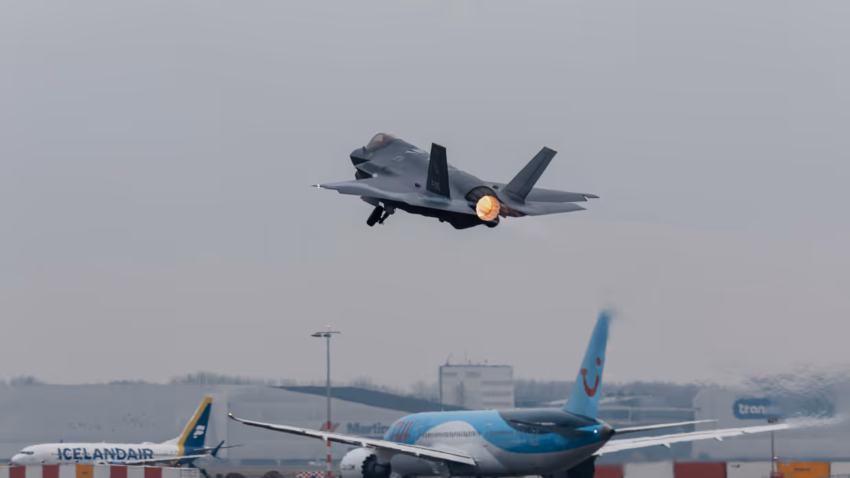 Een f-35 stijgt op vanaf luchthaven Schiphol. Op de achtergrond is een taxiënd vliegtuig van vakantie-aanbieder TUI te zien.