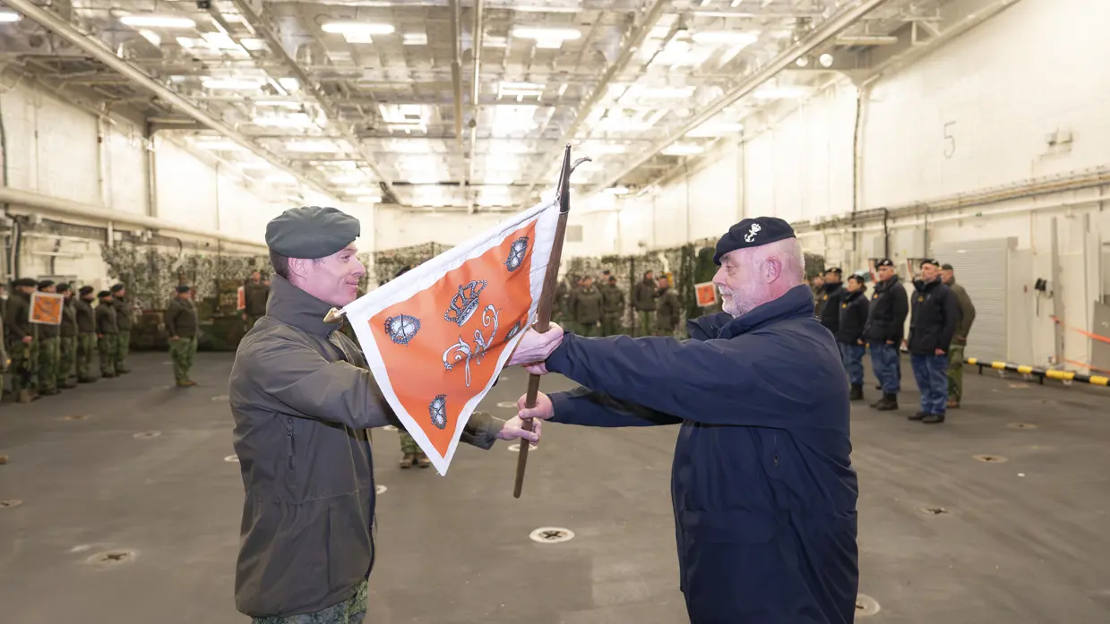 Een militair van de marine krijgt een vlag overhandigd uit handen van een militair van de landmacht.