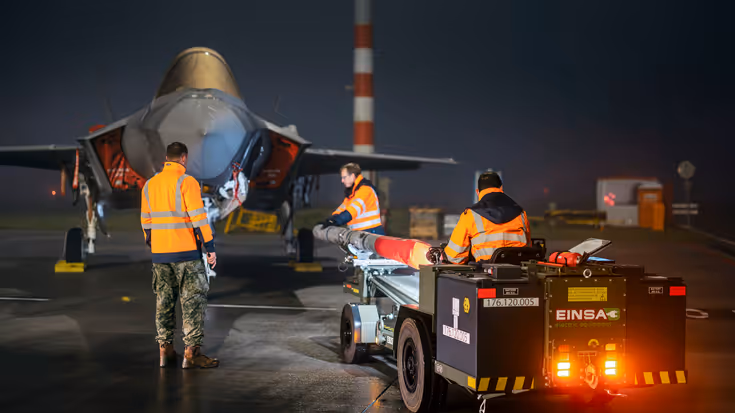 Militairen bewegen met oefenbewapening op een kar naar een geparkeerde F-35. Het is donker, wat een spannend schouwspel creëert.