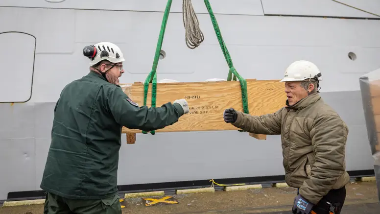 Twee collega’s geven elkaar een boks, met op de achtergrond een houten box, waarop NH90 staat.
