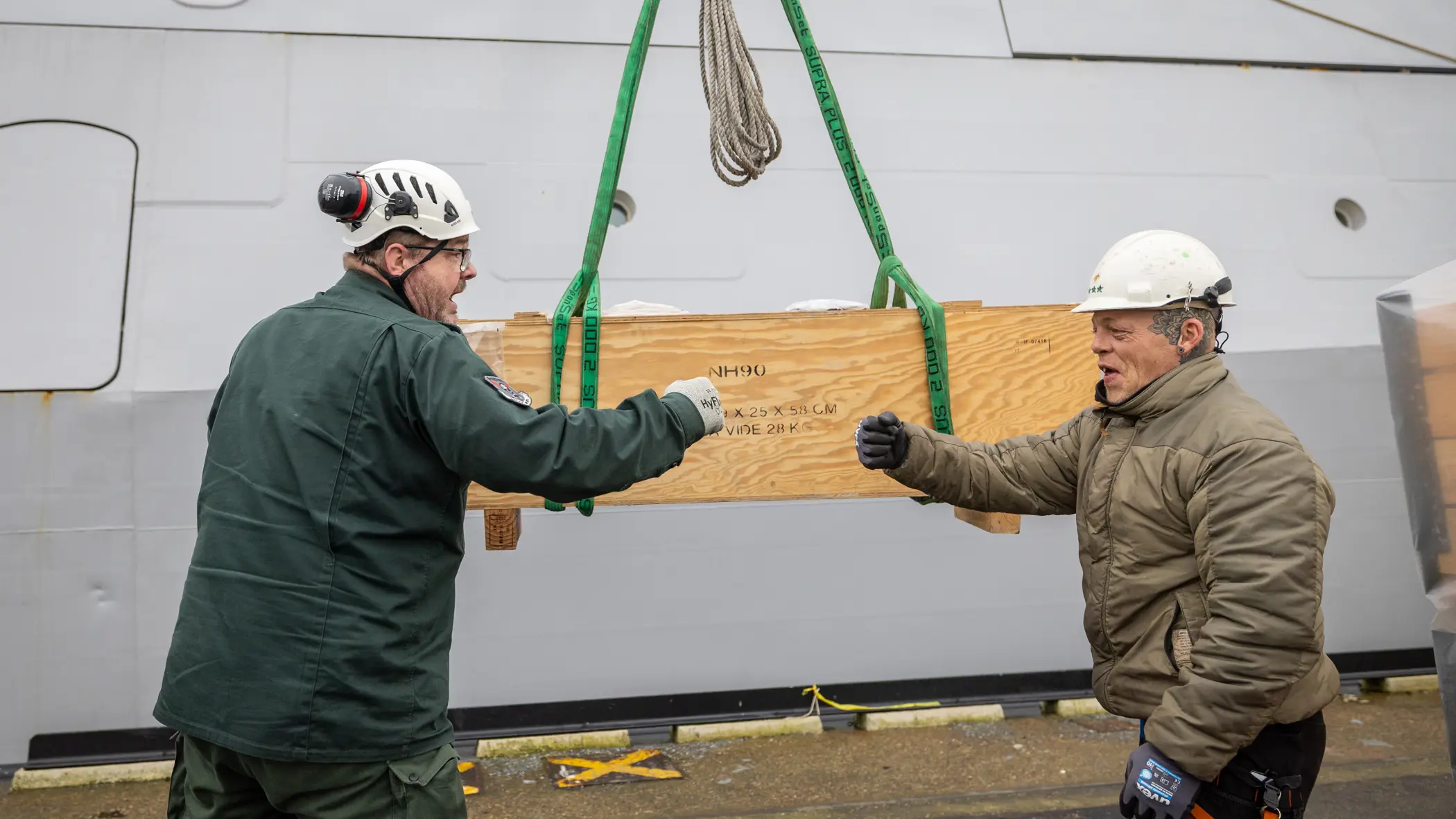 Twee collega’s geven elkaar een boks, met op de achtergrond een houten box, waarop NH90 staat.