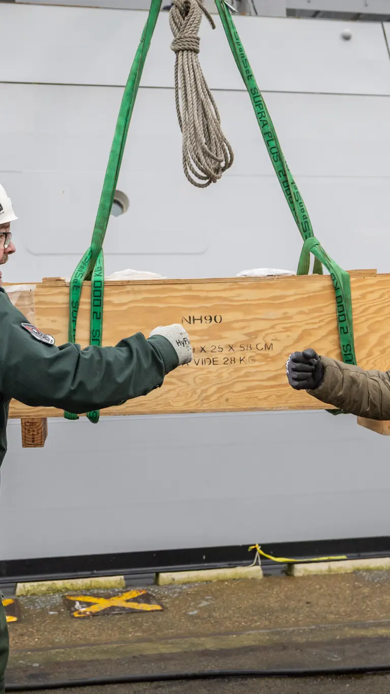 Twee collega’s geven elkaar een boks, met op de achtergrond een houten box, waarop NH90 staat.