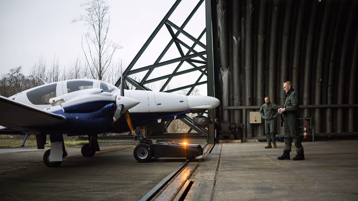 Een patrouillevliegtuig staat links voor de hangar geparkeerd terwijl rechts militairen observeren.
