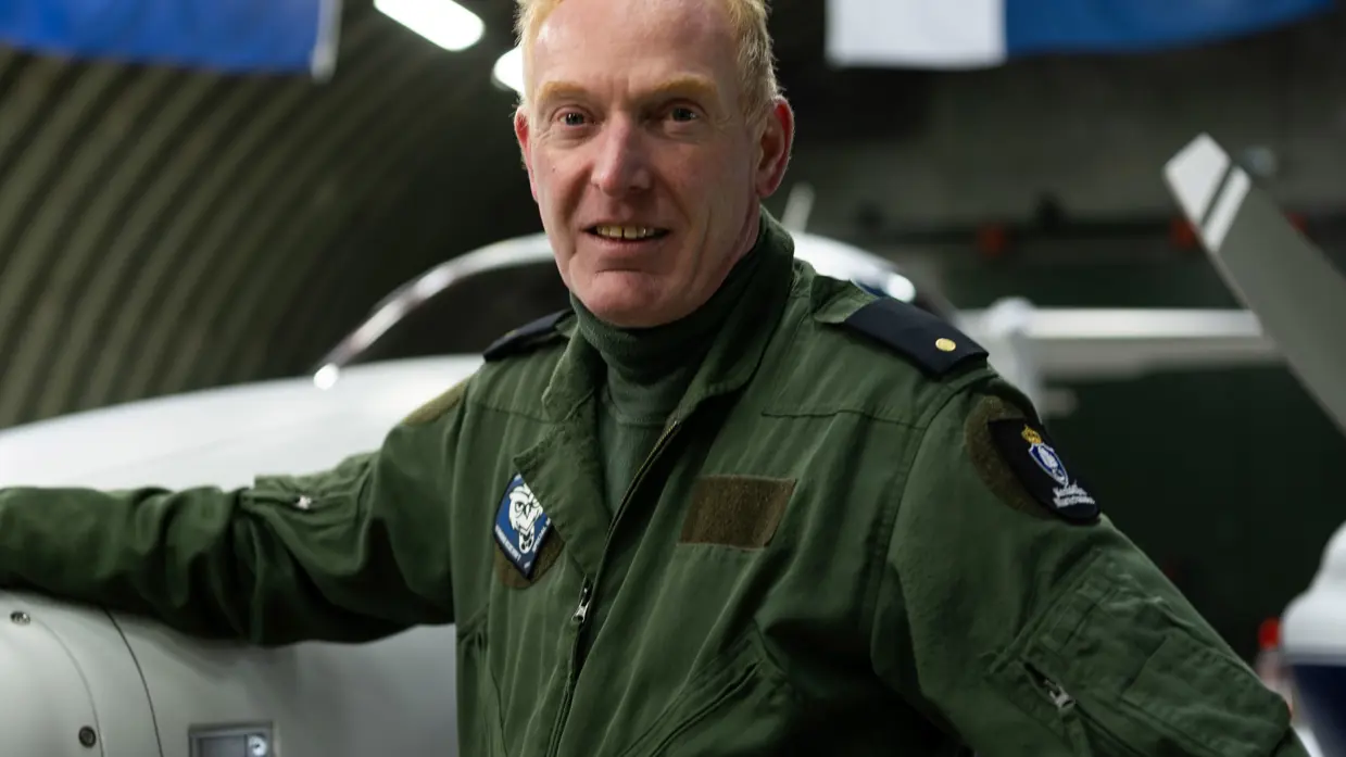 De piloot voor het patrouillevliegtuig poseert naast het vliegtuig in de hangar, vanaf het plafond zijn hangende vlaggen zichtbaar.
