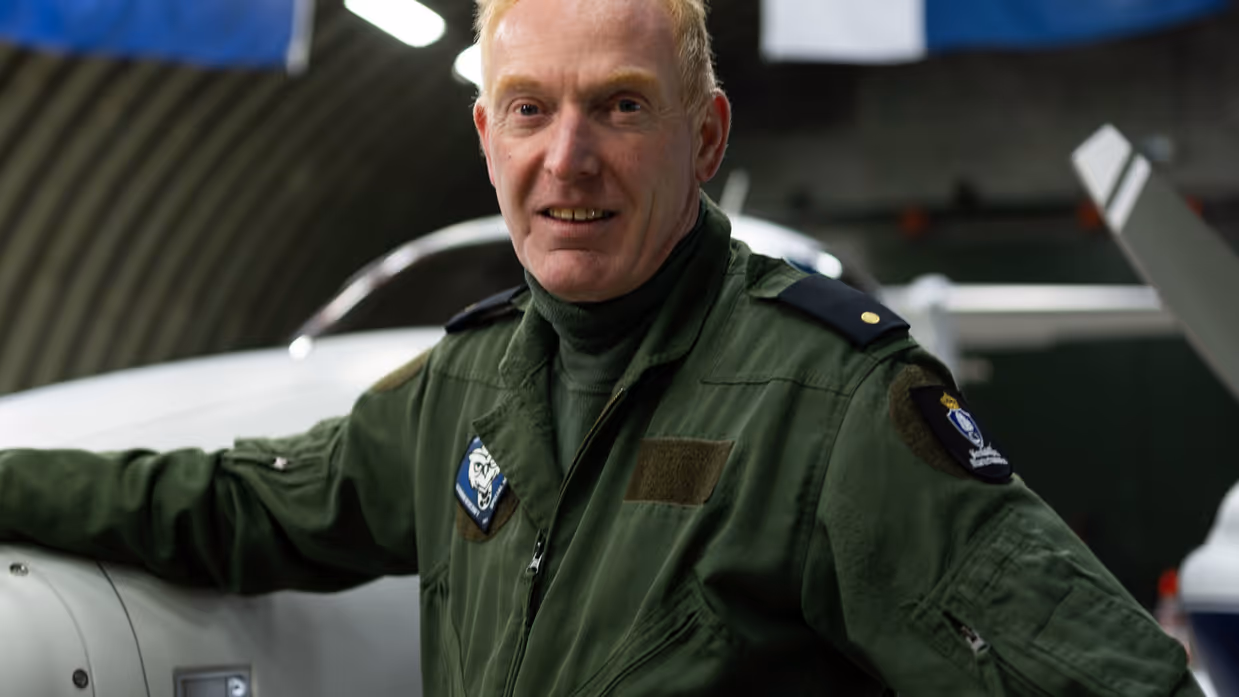 De piloot voor het patrouillevliegtuig poseert naast het vliegtuig in de hangar, vanaf het plafond zijn hangende vlaggen zichtbaar.