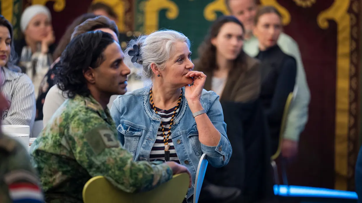 Twee mensen zitten naast elkaar in een zaal: een vrouw met grijs haar en een denim jasje luistert aandachtig, een man met lang haar kijkt mee.