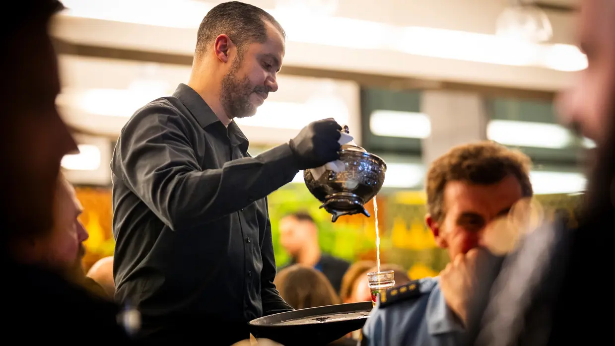 Links: Een man in zwart overhemd schenkt met een glimlach thee uit een traditionele theepot in een klein glas voor een gast, terwijl anderen toekijken. Rechts: Twee mensen zitten naast elkaar in een zaal: een vrouw met grijs haar en een denim jasje luistert aandachtig, een man met lang haar kijkt mee.